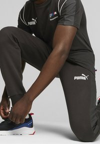 Černý sportovní outfit s bílým logem Puma, skládající se z trička s krátkým rukávem a zúžených kalhot. Tenisky mají modré a červené akcenty.