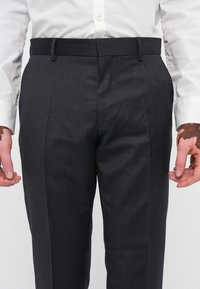 Dunkelgraue, maßgeschneiderte Hose mit glatter Textur, im schmalen Schnitt, mit vorderen Falten und verdecktem Bund. Getragen mit einem weißen Hemd.