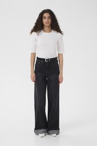 Young woman with curly hair wearing a white short-sleeve top, black wide-leg jeans with cuffs, black belt, and white pointed shoes, standing front.