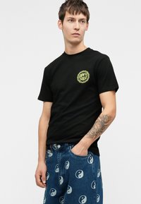 Νεαρός άνδρας που φοράει ένα μαύρο t-shirt Santa Cruz και μπλε τζιν με λευκά σχέδια yin-yang, δείχνοντας τον τατουαρισμένο του βραχίονα, κοιτάζοντας στο πλάι.