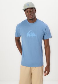 Lyseblå bomulds t-shirt med rund hals, prydet med et bølge-logo i lyseblå. Kombinationen er med khaki shorts og en grå kasket med blå skrift.