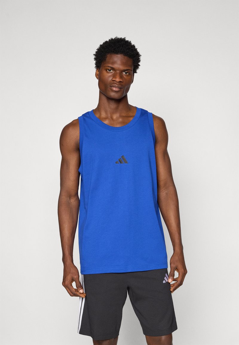 Modra brezrokavna športna majica iz bombaža, z majhnim črnim logotipom Adidas na sredini. Model nosi črne kratke hlače z dodatki.