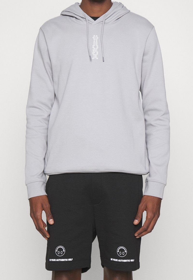 Homme portant un sweat à capuche gris clair et un short noir avec des icônes blanches de casque et le texte « Be Your Authentic Self » sur chaque jambe.