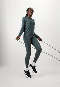 Γυναίκα ντυμένη με αθλητικά ρούχα σε τιρκουάζ, που πηδάει σκοινάκι σε λευκό φόντο, φορώντας μαύρα αθλητικά παπούτσια και λευκές κάλτσες.