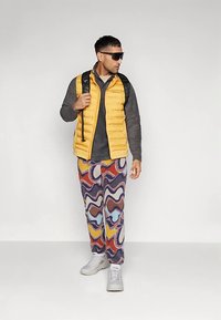 Férfi sárga pufi mellényben, sötét kapucnis pulóverben, színes, absztrakt mintás nadrágban, fehér sportcipőben, napszemüvegben, és egy fekete hátizsákot hordva.