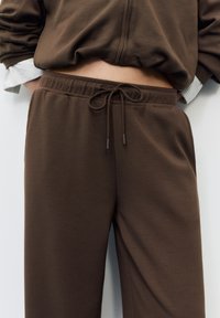 Smeđe trenirke s elastičnim pojasom i bočnim džepovima, izrađene od mekanog, teksturiranog materijala. Imaju opušteni kroj i suptilne detalje šavova.