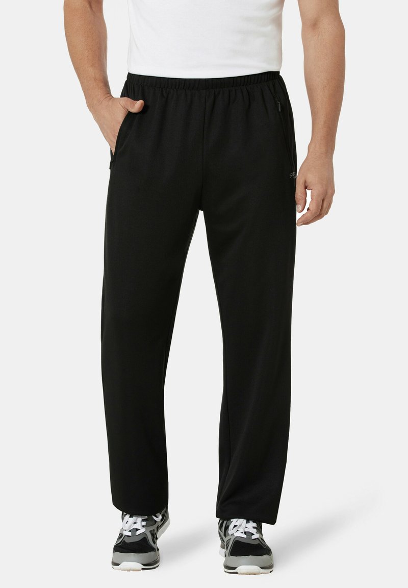 Mann in schwarzen Jogginghosen mit elastischem Bund, schwarzen und weißen Turnschuhen und einem weißen ärmellosen Shirt, mit einer Hand in der Tasche.