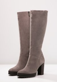 Höga grå mockastövlar för kvinnor med sidodragkedja och klackar, placerade bredvid varandra på en vit yta.