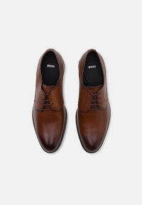 Zapatos de vestir de cuero marrón con un acabado suave, que presentan un sistema de cordones cerrado, punta redondeada e interior negro con el logo de la marca.