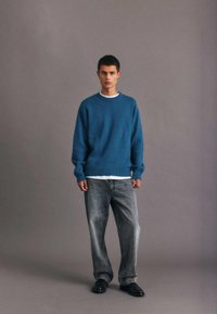 Suéter azul con puños y dobladillo acanalados, superpuesto a una camiseta blanca. Un par de jeans grises holgados y zapatos negros, de pie frente a un fondo gris.