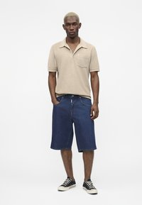 Homme aux cheveux blonds courts se tenant les mains dans les poches, portant un polo beige à manches courtes, un short en jean longueur genoux et des baskets noires.