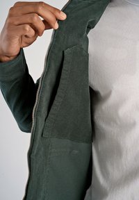 Groene jacks met ritssluiting en een zachte textuur, met een binnenzak aan de linkerzijde, gedragen over een lichtgrijze t-shirt.