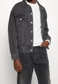 Jeansjacka i blekt svart med framfickor och silvriga knappar, lager på en vit ribbad tröja, i kombination med matchande svarta jeans.