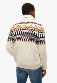 Suéter de punto crema con patrones geométricos en burdeos, negro y amarillo. Presenta un cuello alto y puños de canalé.