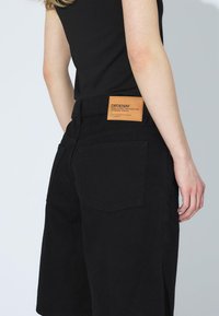 Los pantalones cortos de mezclilla negra tienen una cintura alta, cinco bolsillos y una etiqueta de cuero marrón en la parte posterior con los detalles de la marca. Textura suave, corte recto.