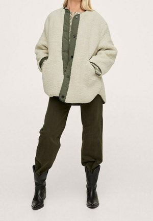 Frau steht und trägt eine hellbeige, übergroße Fleecejacke mit grünem Knopfleistenstreifen, dunkelgrüne Hose und schwarze Stiefel bis zur Mitte der Wade.
