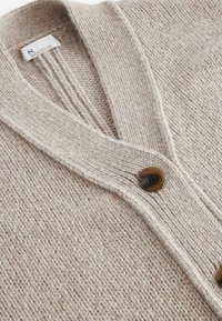 Cardigan beige en maille épaisse avec col châle et grands boutons marron, montrant un gros plan de la texture et de l'étiquette à l'intérieur du col.