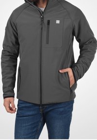 Mann in einer dunkelgrauen Zip-Jacke mit vertikeller Brusttasche und Händen in den Taschen, kombiniert mit blauen Jeans.