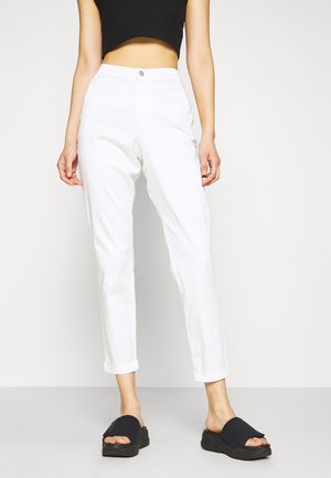 Białe spodnie o prostych nogawkach z wysokim stanem, zapięciem na guziki i podwiniętymi mankietami. W duecie z czarnymi sandałami wsuwanymi na neutralnym tle.