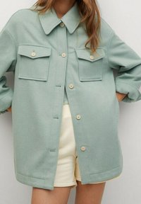 Giacca camicia oversized in misto lana verde chiaro con colletto a punta, due tasche sul petto e chiusura a bottoni, caratterizzata da una texture morbida.