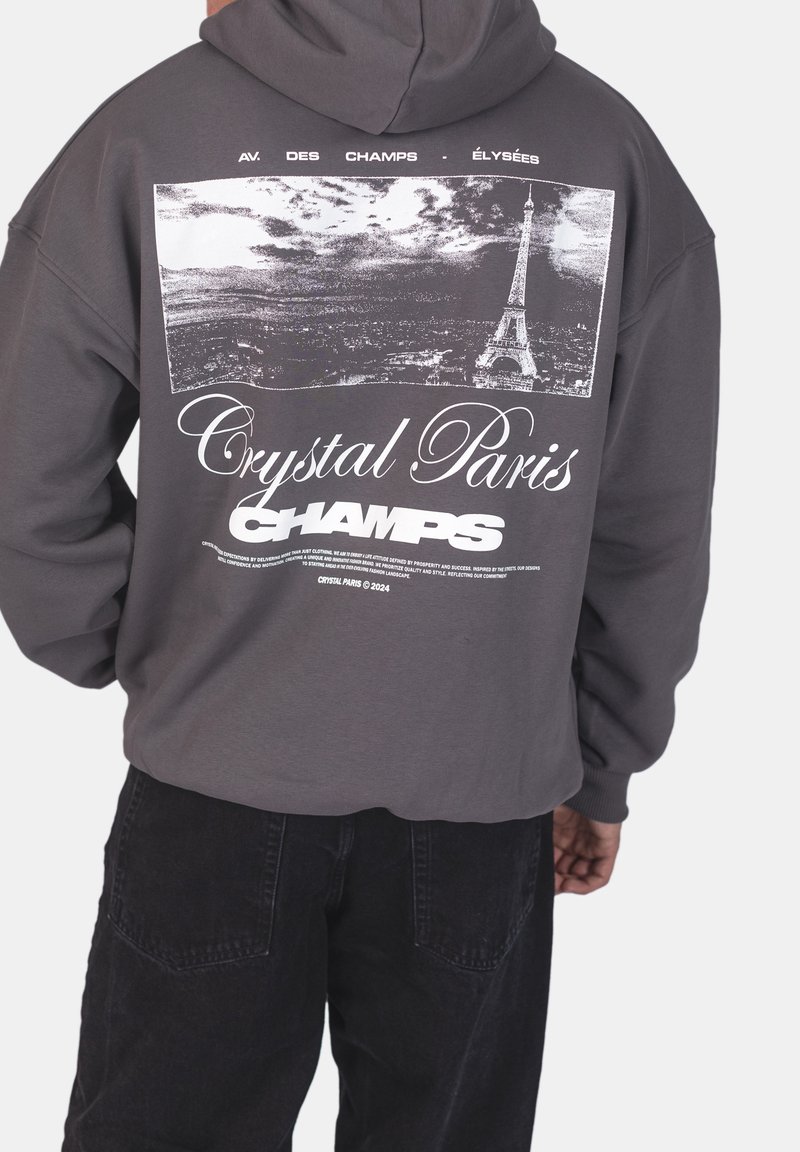 Personne portant un sweat à capuche sombre avec un motif noir et blanc de la Tour Eiffel et de la silhouette de la ville avec le texte "Crystal Paris Champs" dans le dos.