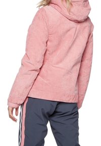 Rosa Cordjacke mit Kapuze, die eine weiche Textur, ein geripptes Muster und einen geraden Schnitt aufweist, kombiniert mit dunklen Hosen mit seitlichen Streifen.