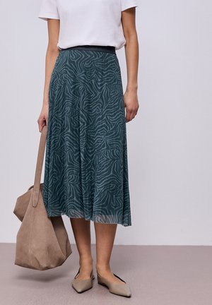 Femme portant une jupe midi turquoise à imprimé zèbre abstrait, un haut blanc à manches courtes, des ballerines beiges et tenant un grand sac fourre-tout beige.