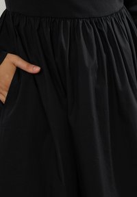 Henkilö, joka on pukeutunut kerättyyn mustaan hameeseen, vasen käsi on puvun taskussa.