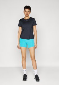 Czarny sportowy top z krótkim rękawem z logo Nike, w połączeniu z dopasowanymi turkusowymi szortami. Modelka ma na sobie czarne trampki i białe skarpetki.