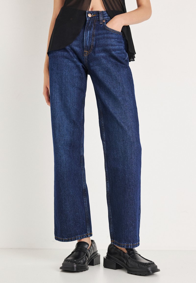 Augsta vidukļa zilas džinsa bikses ar taisnu kāju piegriezumu, kurās ir oranžas diegu šuves un priekšējā kabata. Kopā ar melnām loafer tipa kurpēm.