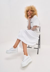 Baskets platform blanches avec une texture lisse, présentant des accents gris minimalistes et un design à lacets. Portées avec une longue jupe en satin blanche.