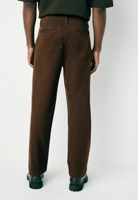 Person wearing dark brown straight-leg pants and black chunky shoes, standing against a plain light background, viewed from behind.