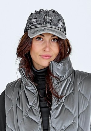 Frau mit braunen Haaren trägt eine abgenutzte graue Jeanskappe, einen schwarzen Rollkragenpullover und eine glänzende graue gesteppte Daunenweste mit hohem Kragen.