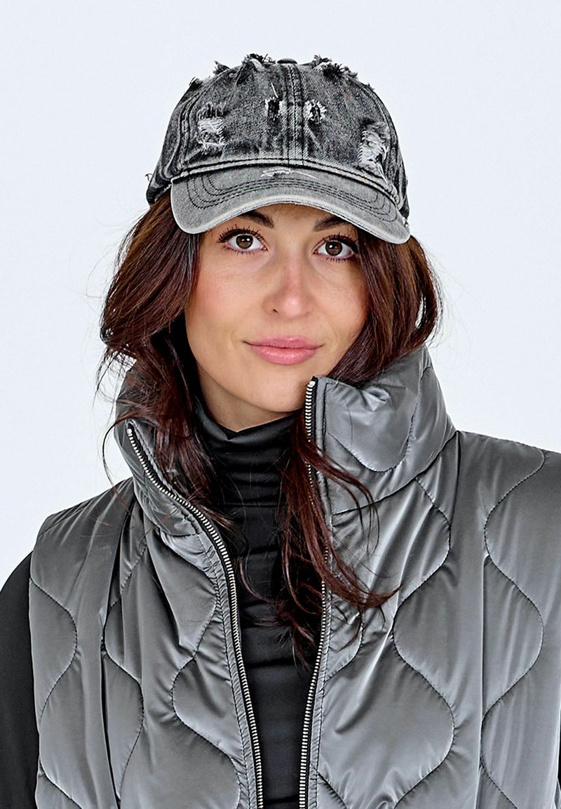 Femme aux cheveux bruns portant une casquette en denim gris usée, un col roulé noir et un gilet matelassé gris brillant avec col montant.