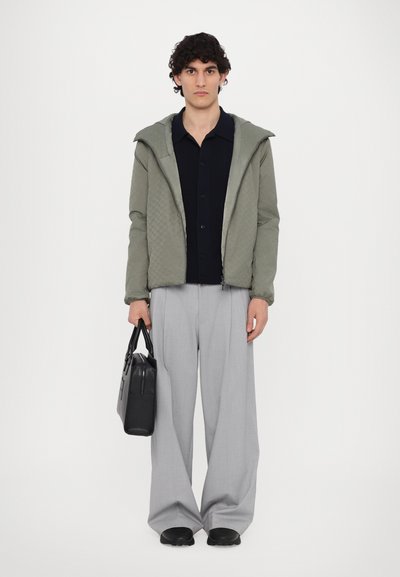 Vadderad, vadderad grön jacka med huva, tillsammans med en mörk knapprad skjorta och ljusgrå vidbyxor. Håller en svart väska.