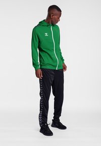 Grøn zip-up hoodie med hvid lynlås og logo. Sorte bukser med grå chevronmønster på siderne. Båret med sorte træningssko.