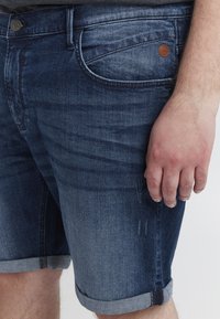 Jeans kratke hlače v temno modri barvi z zbledelim videzom. Imajo zavite robove in rjavo oznako na sprednji žepu. Poudarjen pogled na rob in roko.