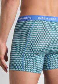 Modra elastična višina pasu s tekstom "BJÖRN BORG"; zelena in modra geometrijska diamantska zasnova; prilegajoč se raztegljiv material; dolžina do sredine stegen.