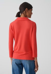 Koraal gebreide top met een kraag en driekwart mouwen, gecombineerd met lichtblauwe spijkerbroek. Zachte textuur, relaxte pasvorm en eenvoudig ontwerp.