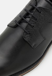 Scarpa in pelle nera con finitura liscia, caratterizzata da dettagli testurizzati e sei asole per i lacci. Punta arrotondata e cuciture a contrasto.