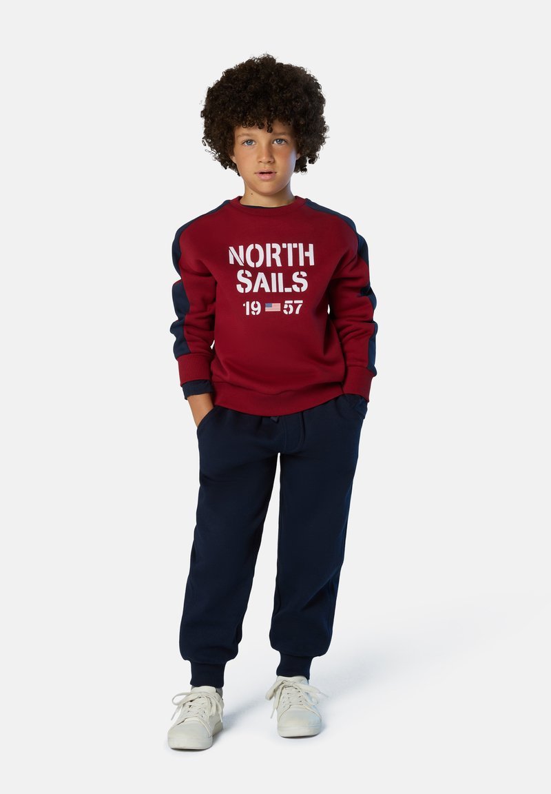 Felpa color bordeaux con la scritta "NORTH SAILS 1957", dettagli blu navy sulle maniche, abbinata a pantaloni della tuta blu navy. Sneakers bianche completano il look.