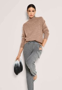 Frau mit dunklen Haaren, die einen braunen Rollkragenpullover und graue Cargo-Hosen trägt, hält eine schwarz-weiße Fransen-Clutch und posiert mit der Hand in der Tasche.