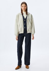 Felpa grigia chiara con cappuccio e tasche frontali, abbinata a una tuta in denim scuro con bottoni dorati e pantaloni a gamba larga. Stivaletti marroni alla caviglia.