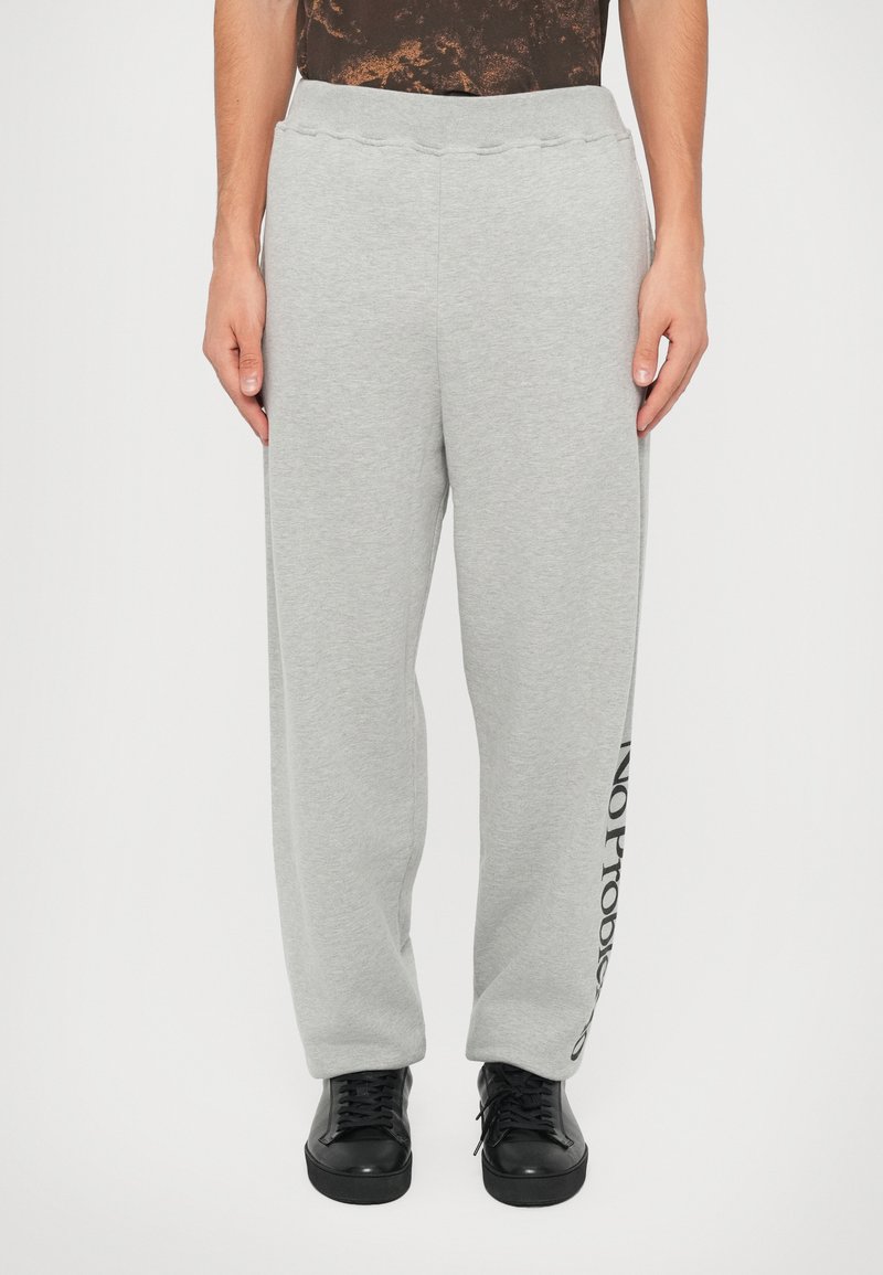 Pantalones de chándal grises con un ajuste holgado, cintura elástica y detalle impreso de "NO PROBLEM" en negro a lo largo de la pierna izquierda. Textura suave y lisa.
