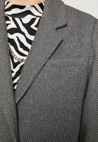 Blazer gris de espiga con solapas con muesca y hombros estructurados, superpuesto a una blusa de patrón de cebra en blanco y negro y un collar de cadena.