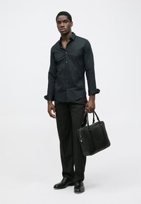 Μαύρο πουκάμισο με γιακά, συνδυασμένο με μαύρο παντελόνι φόρεμα και στιλβωμένα μαύρα παπούτσια. Κρατά μια μαύρη δερμάτινη χαρτοφύλακα.