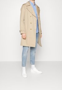 Beige trench coat, jossa on kaksinkertainen napitus, kultaiset napit ja leveä kaulus, vaaleansinisen paidan päällä, yhdistettynä vaaleisiin farkkuihin ja valkoisiin kenkiin.