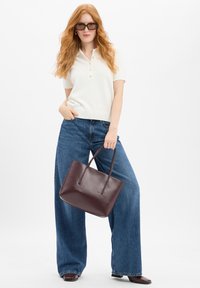 Bordeaux leren totebag met een gladde textuur en een eenvoudig ontwerp, vastgehouden door een persoon in een crèmekleurig polo-shirt en brede blauwe jeans.