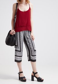 Rotes ärmelloses Oberteil, gemusterte schwarz-weiße Culottes, schwarze Plattformsandalen und eine gesteppten schwarzen Handtasche. Einfaches und modernes Design.
