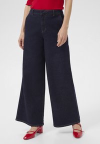 Löysät tummansiniset farkut korkealla vyötäröllä, joissa on näkyvät ompelusyksityiskohdat. Yhdistetty kiiltäviin punaisiin kenkiin.